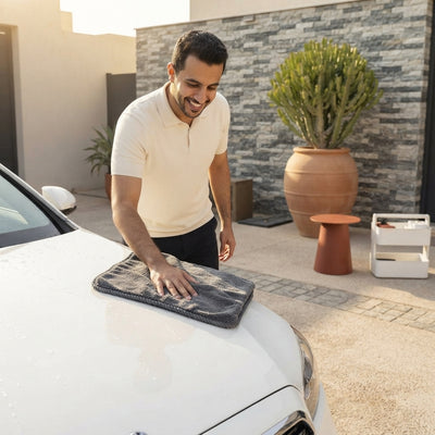 Saudi man using a grey microfiber cloth to clean a car in a sunny, modern driveway.